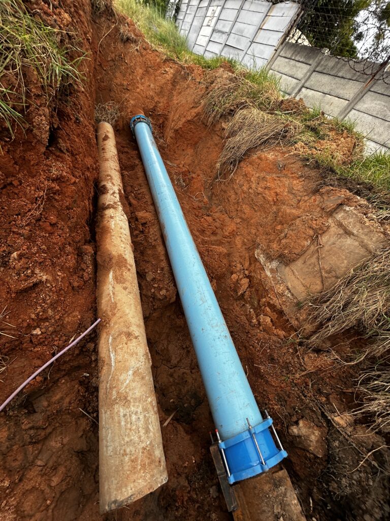 Two large water pipes—old concrete and new blue PVC—exposed in a deeply excavated trench of red earth, connected with a mechanical coupling during an emergency repair project by Saunders Plumbing.