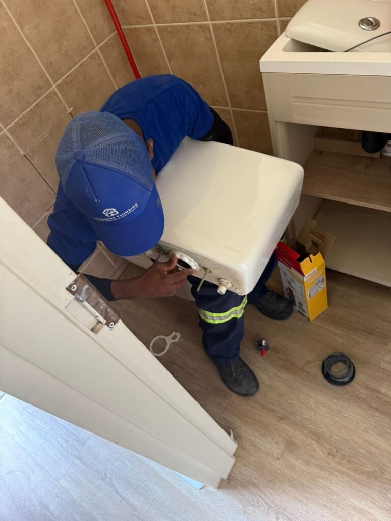 A Saunders Plumbing professional in branded safety gear kneeling to perform a toilet repair or installation in a neat bathroom.